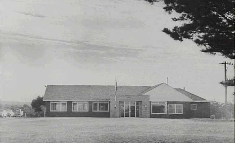 Black and white photo of a large single-story building with a gabled roof, situated on a grassy field with a tree partially visible on the right and power lines in the background.