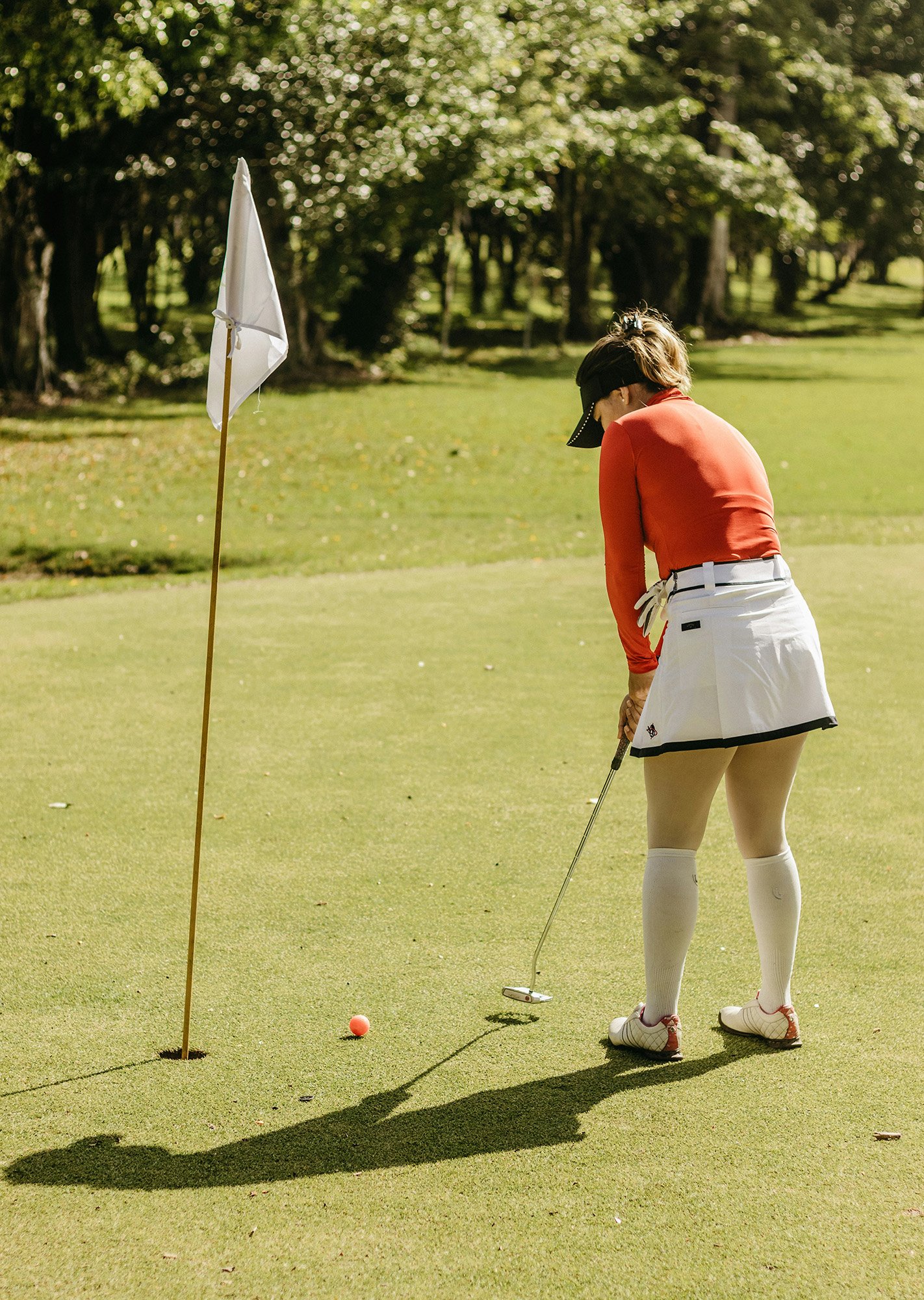 A woman in a red long-sleeve shirt, white skirt, white knee-high socks, and cap putting a golf ball on the green with a putter, near a white flagstick on a golf course during daytime.