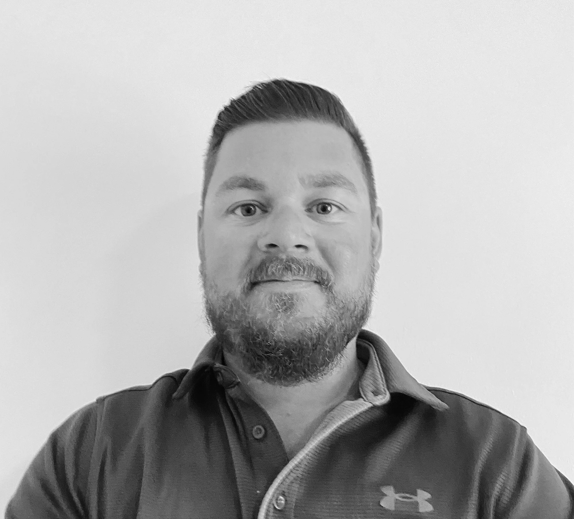 Black and white photo of a man with short dark hair and a beard, wearing a dark polo shirt with a logo on it, standing against a plain background.