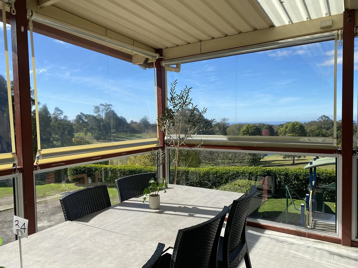 Outdoor patio with a table, chairs, and a potted plant, overlooking a scenic landscape with grass, trees, and a blue sky.
