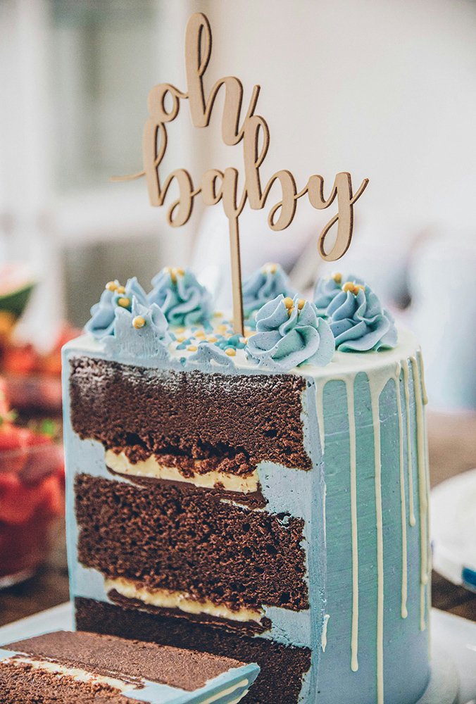 A slice of cake with three layers of chocolate cake and cream filling, decorated with blue frosting and yellow sprinkles, topped with a wooden 'oh baby' cake topper.