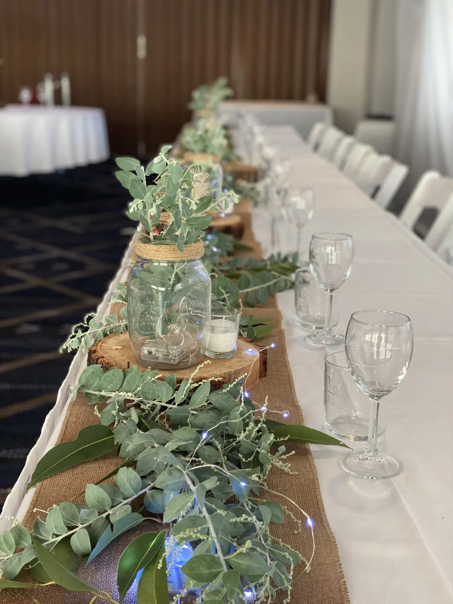 A long banquet table decorated with greenery in mason jars, wood slices, and string lights, set for a celebration or event.