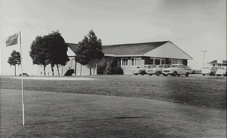 Black and white photo of a golf course with a flag in the foreground, a clubhouse with trees, and parked cars in the background.