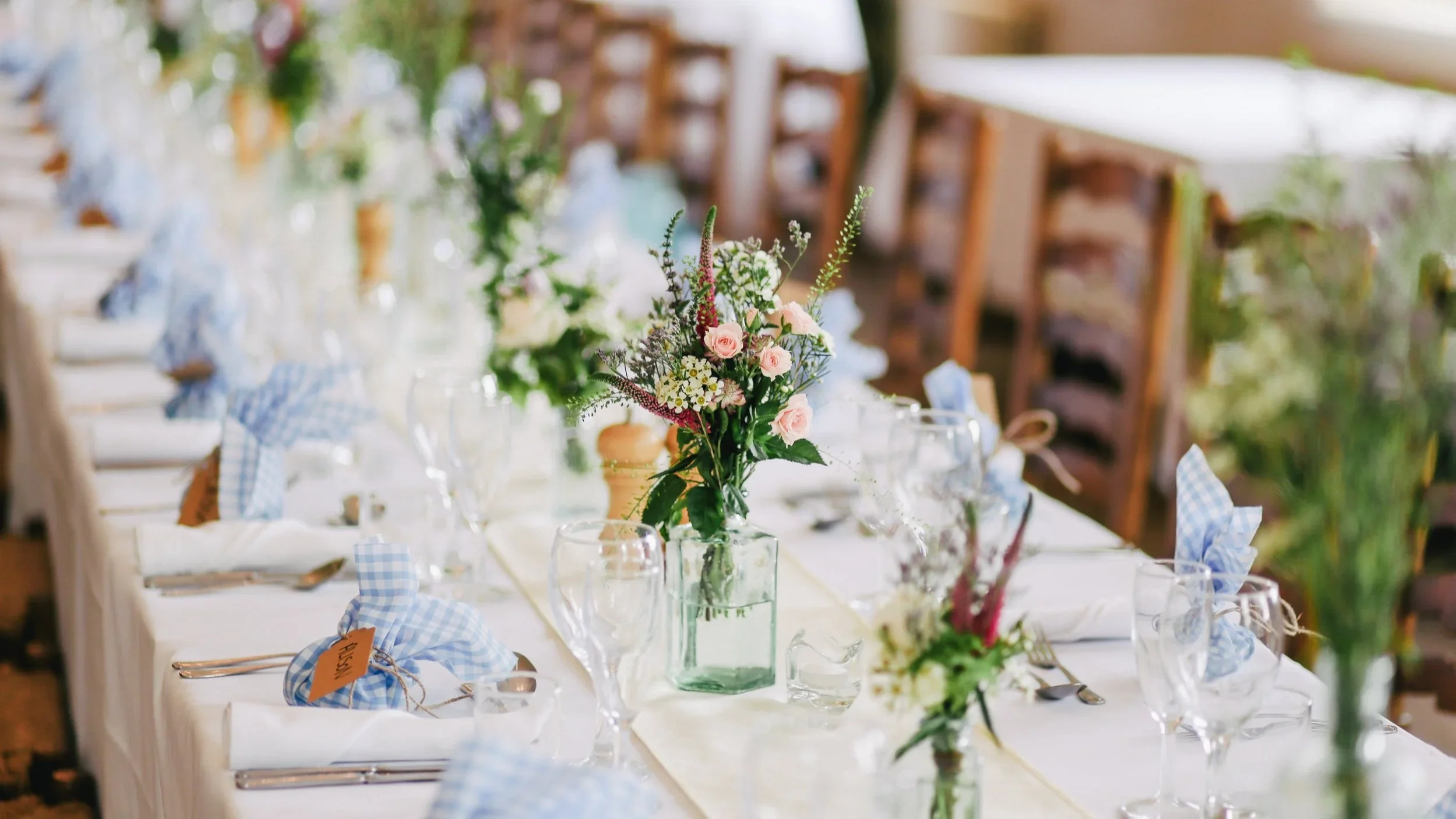 Elegant banquet table set with white tablecloth, floral centerpieces in glass vases, blue checked napkins, and glassware at a rustic venue.