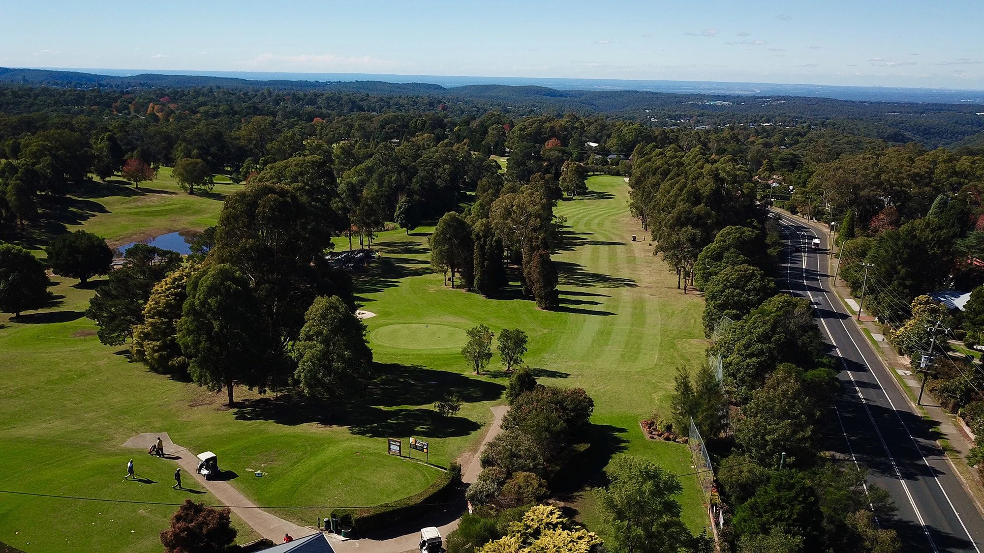 Aerial view of a golf course with green fairways, trees, a small pond, and a golf hole. A road runs alongside the course on the right, with houses and trees lining the street. The landscape extends to distant hills and a blue sky with some clouds.
