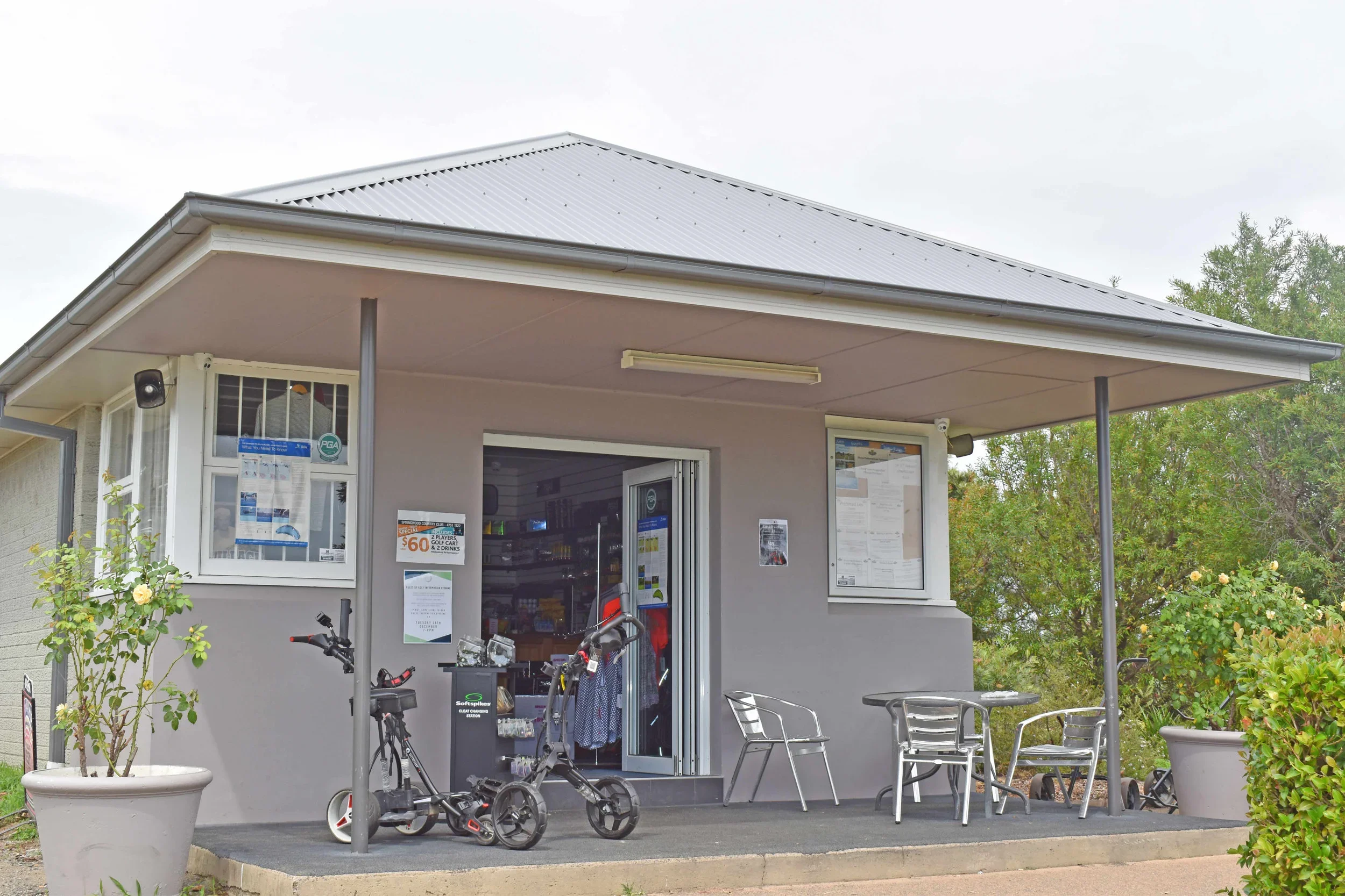 Front view of the pro shop with aluminum tables and chairs outside, two bicycles parked in front, and greenery surrounding the building.