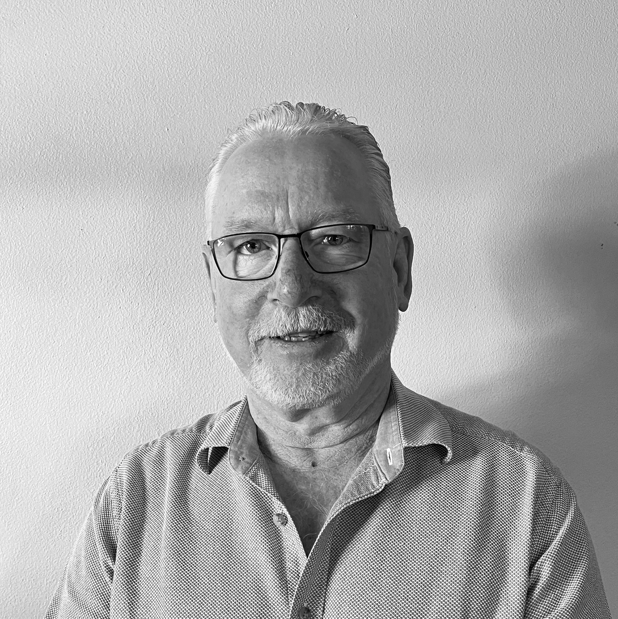 Black and white photo of a smiling man with glasses, gray hair, and a beard, wearing a collared shirt, standing against a plain wall.