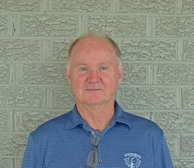 A middle-aged man with short, light-colored hair, wearing a blue polo shirt with a logo that reads 'Springwood Country Club,' standing against a textured gray brick wall.