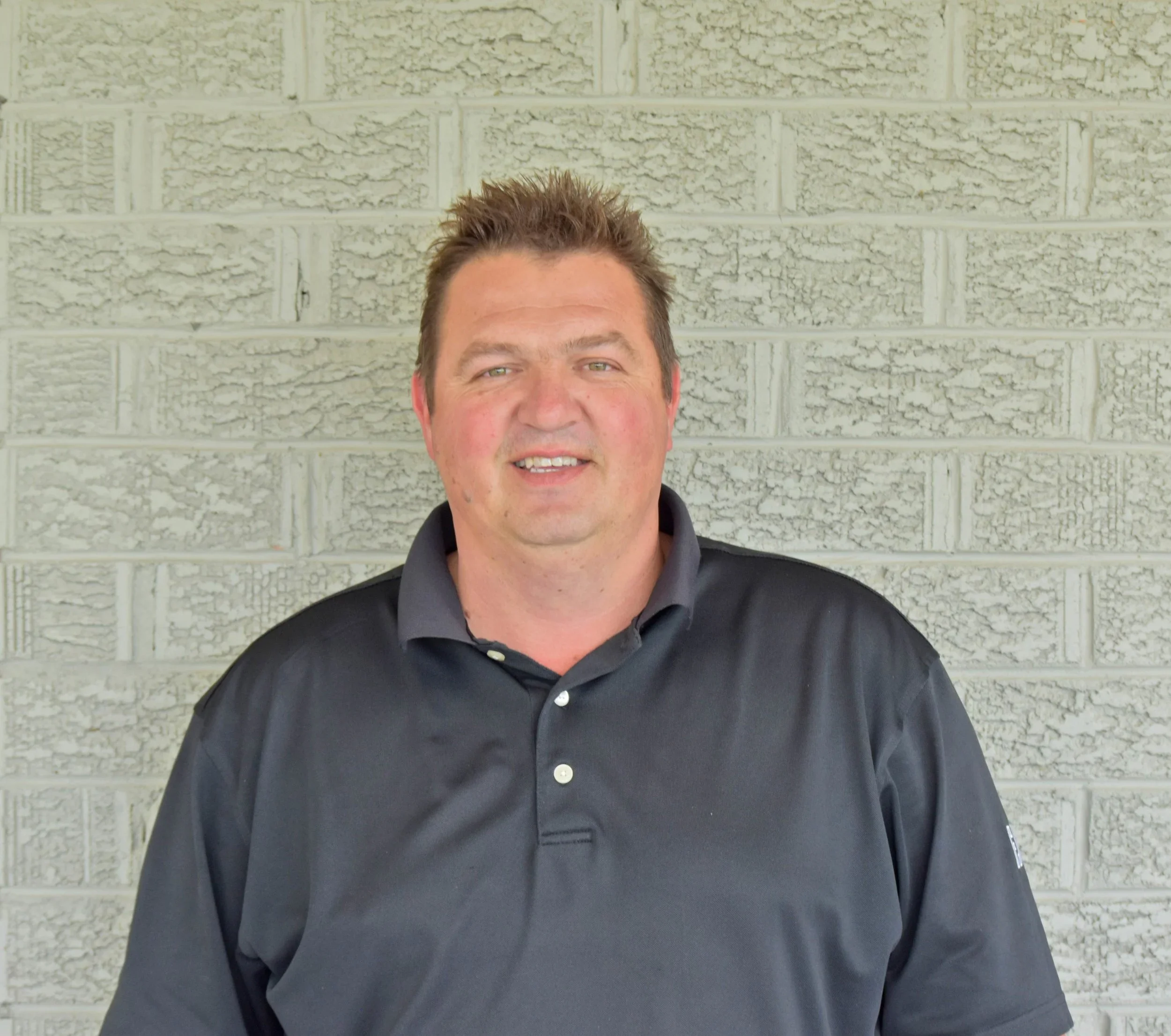 A man standing in front of a light gray brick wall, wearing a black polo shirt.