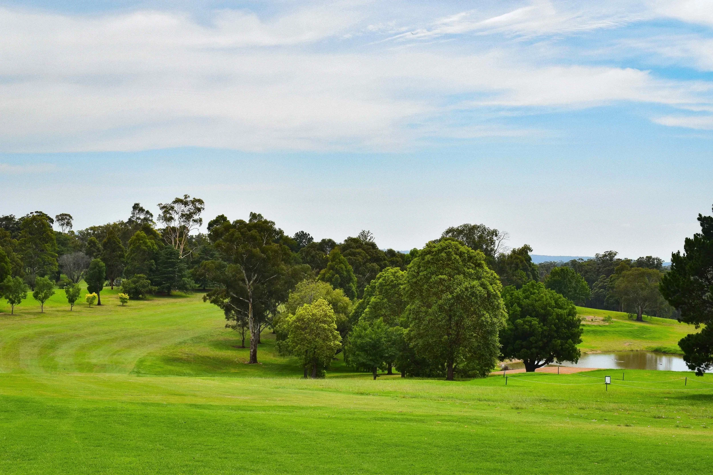 A lush green golf course with many trees and a small pond in the background under a partly cloudy sky.