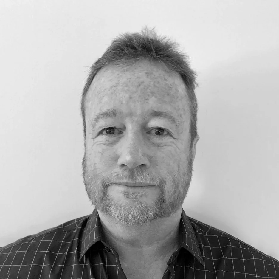 Black and white portrait of a man with short, slightly disheveled hair and a beard, wearing a checkered shirt, standing against a plain white background.