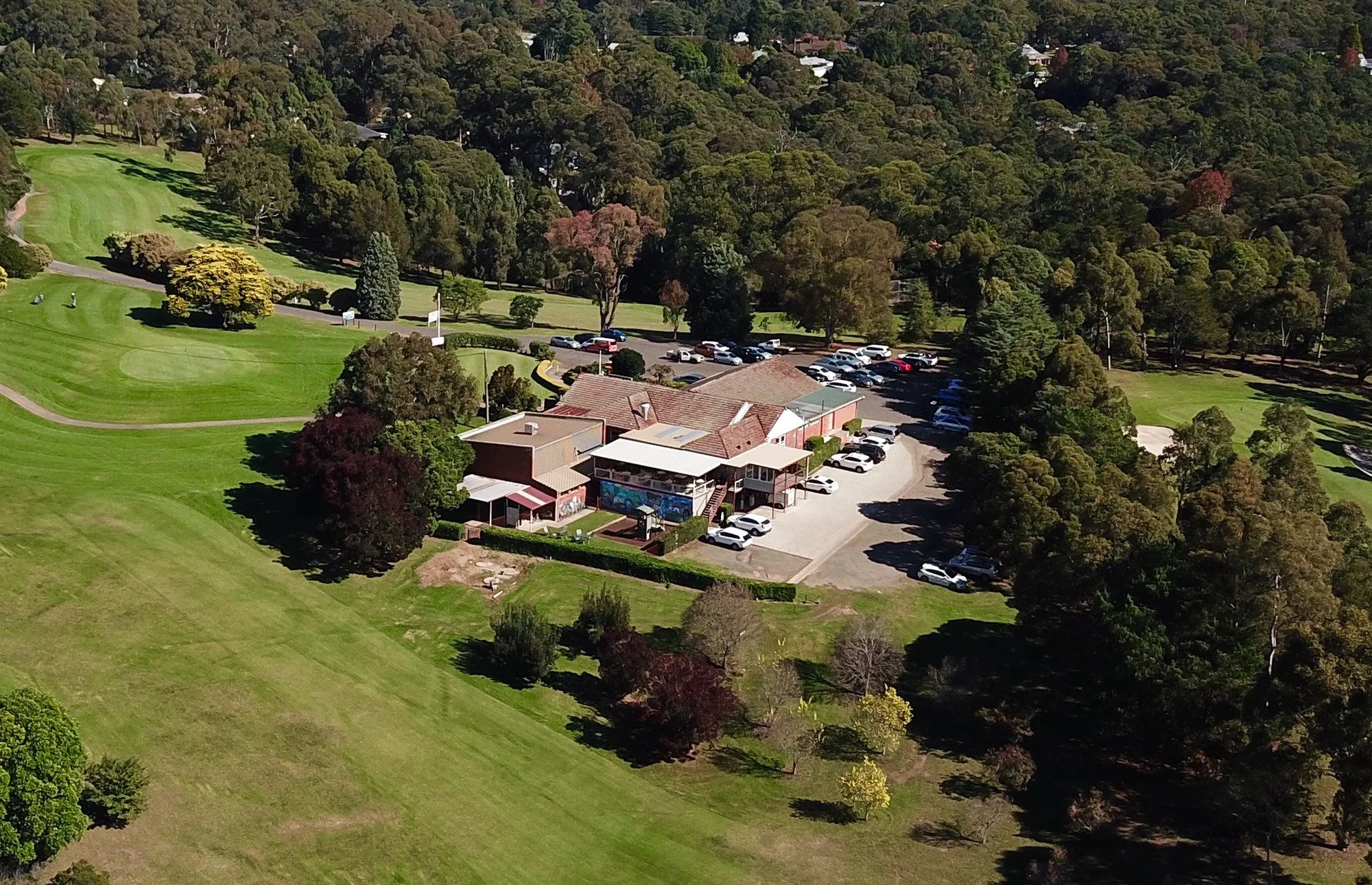 An aerial view of a clubhouse surrounded by a parking lot, trees, and a golf course.