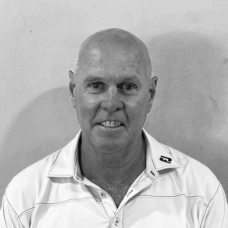 A black and white photo of a middle-aged man with a shaved head, wearing a collared shirt with a logo on the collar, smiling slightly while looking at the camera.