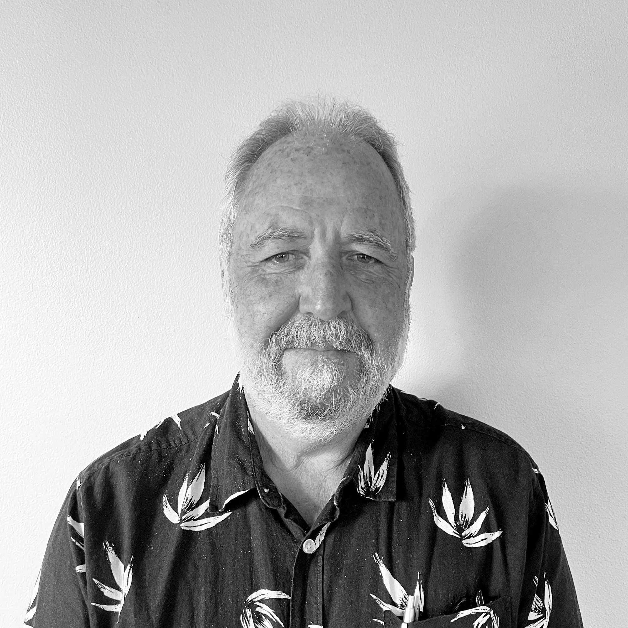 Black and white portrait of an older man with gray hair and beard, wearing a patterned button-up shirt, standing against a plain background.
