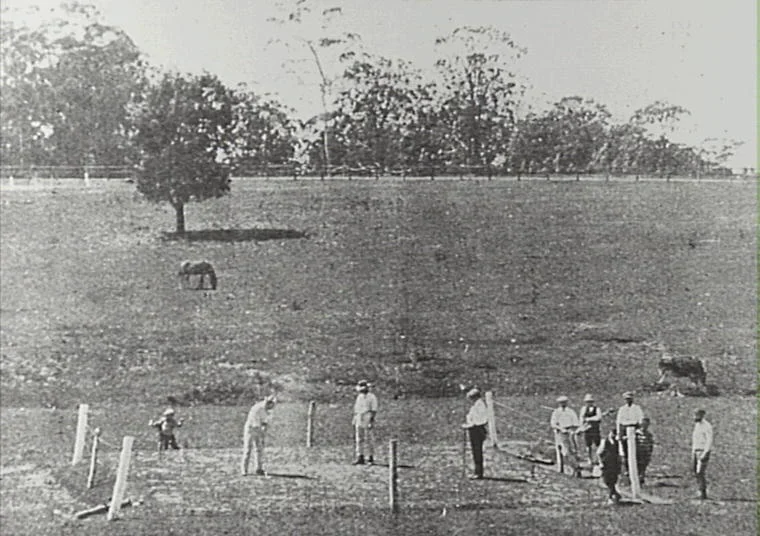 A group of men and a boy in early 20th-century clothing standing and working on a small fenced area on a grassy field, with trees and a horse in the background.