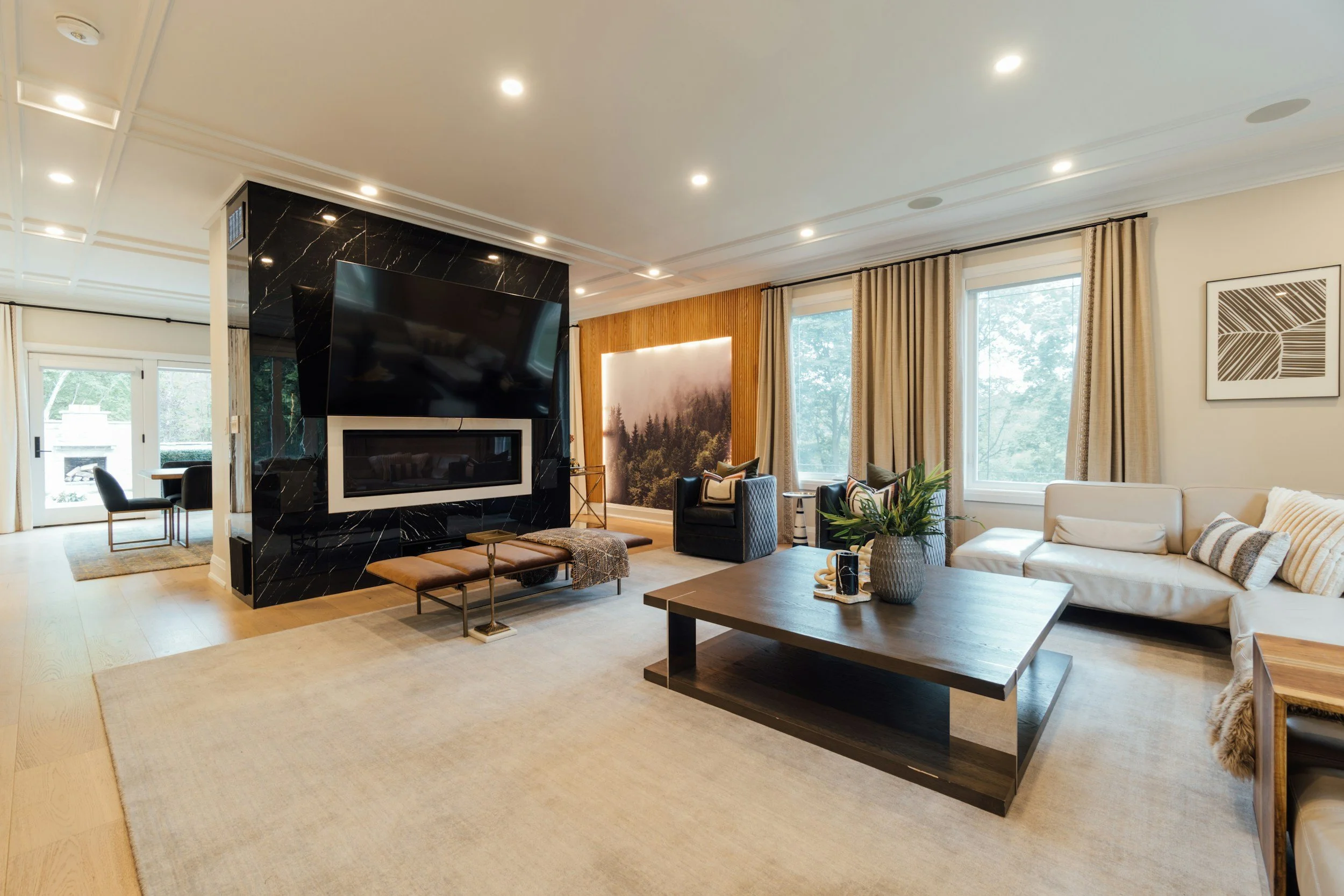 Living room with a black marble fireplace, large TV, beige sofa, black armchairs, coffee table, and large windows with beige curtains, decorated with framed art and green plants. Houston, TX interior design, Room furnishing packages.  Room makeovers.
