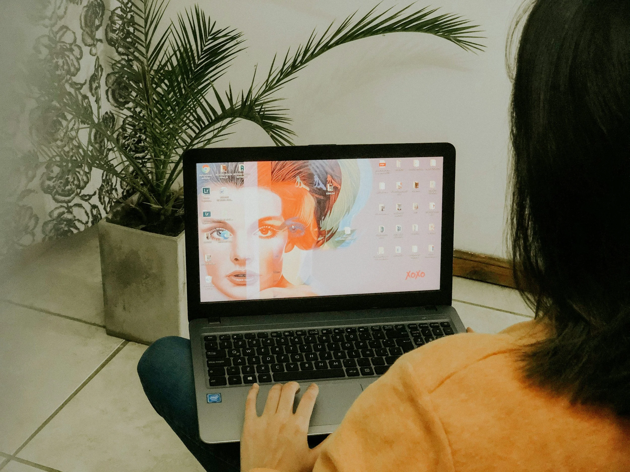 A person with dark hair working on a laptop with a colorful abstract art wallpaper of a woman's face. There is a potted plant with long, green leaves on the left side of the image. The workspace is on a tiled floor, and the person is sitting with their right hand on the keyboard.