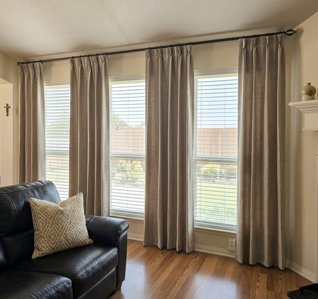 Living room with three large windows covered with beige curtains, a black leather sofa with a beige patterned pillow, and hardwood flooring. Houston TX