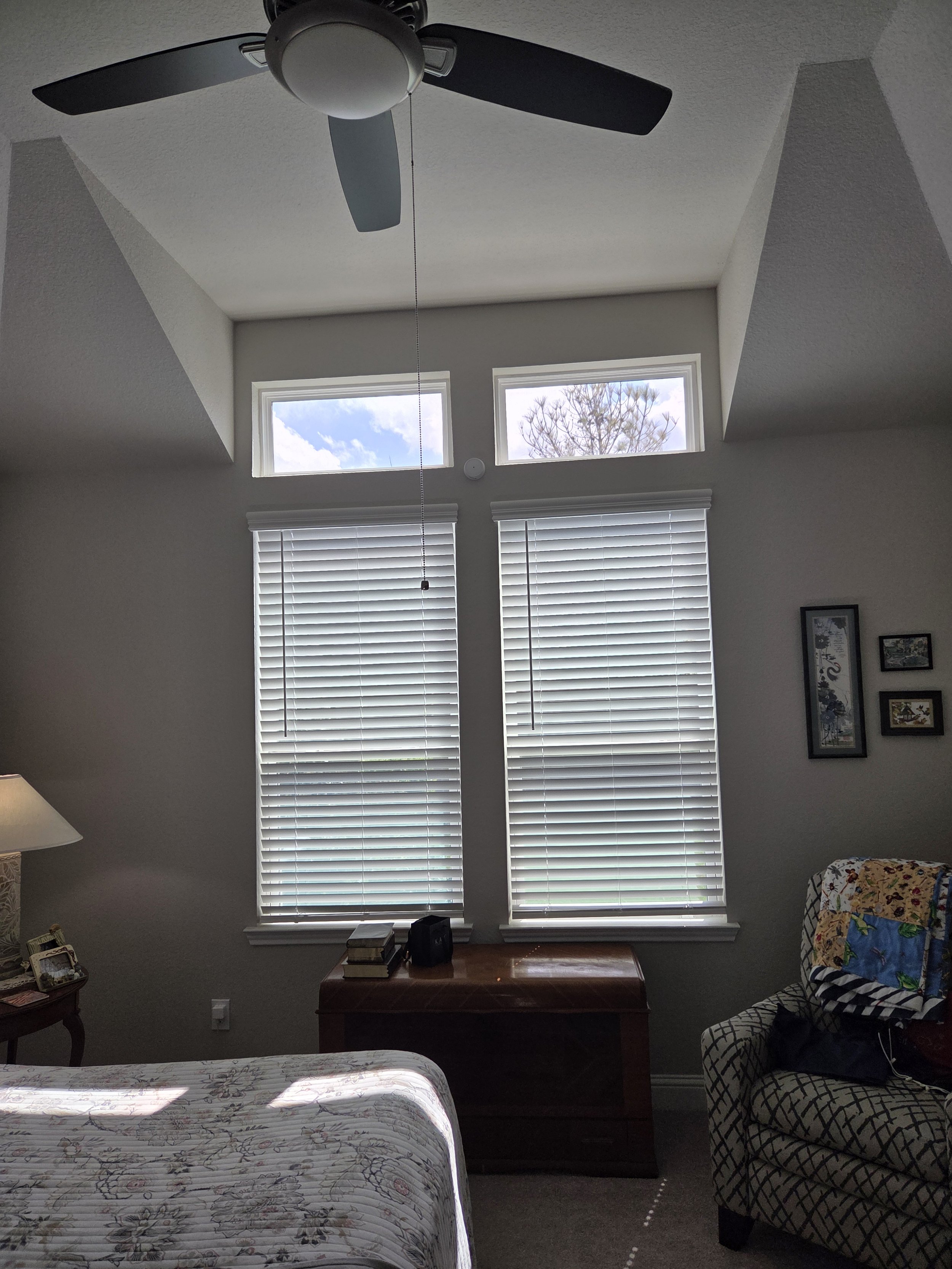 A bedroom with a window wall, covered with white blinds, and a ceiling fan. There is a dresser under the window, a chair with a patterned cover, and a bed with a floral quilt.