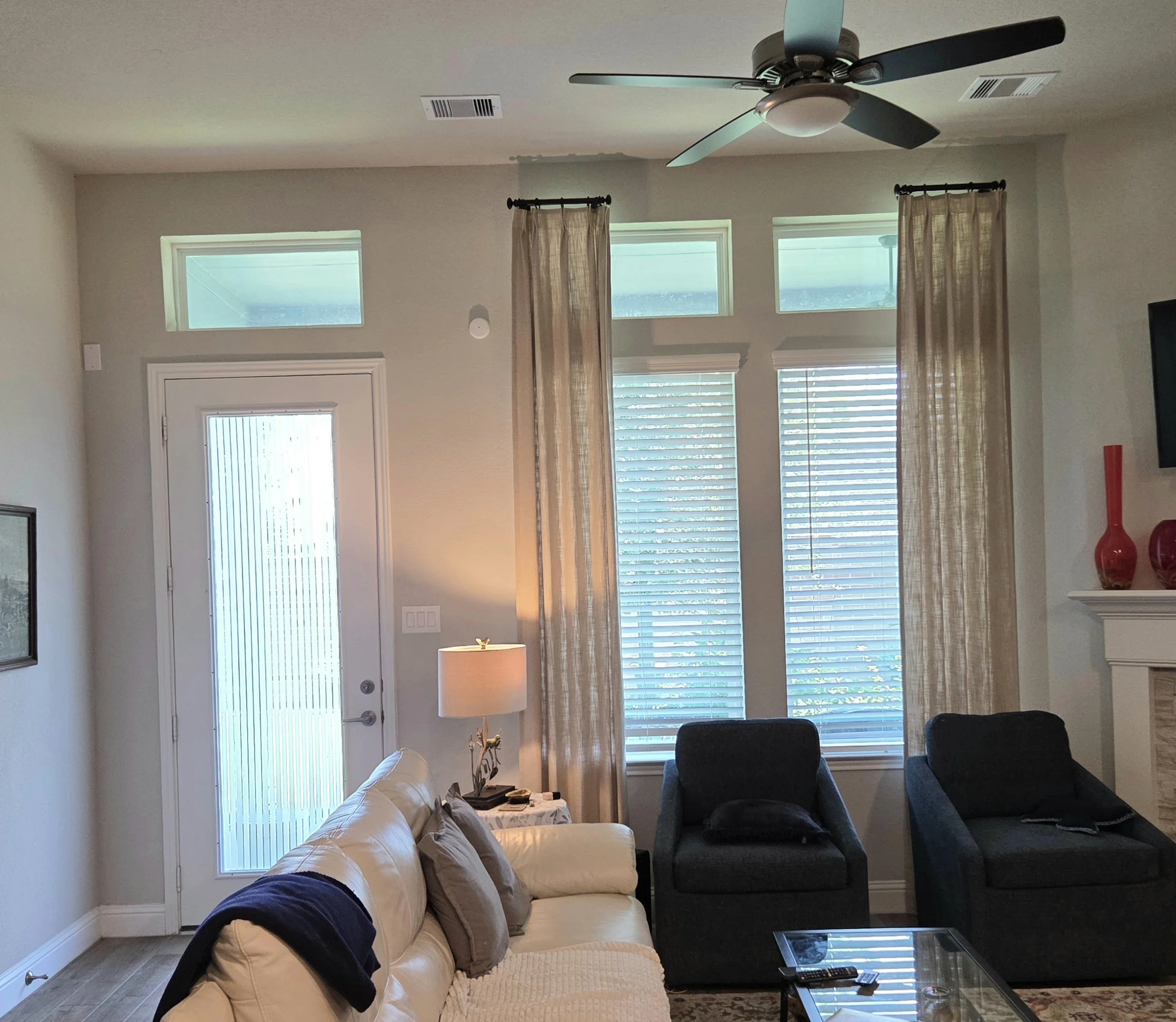 Living room with beige couch, dark blue armchair, glass-top coffee table, and windows with blinds and custom designed light curtains, lit by natural light from outside.