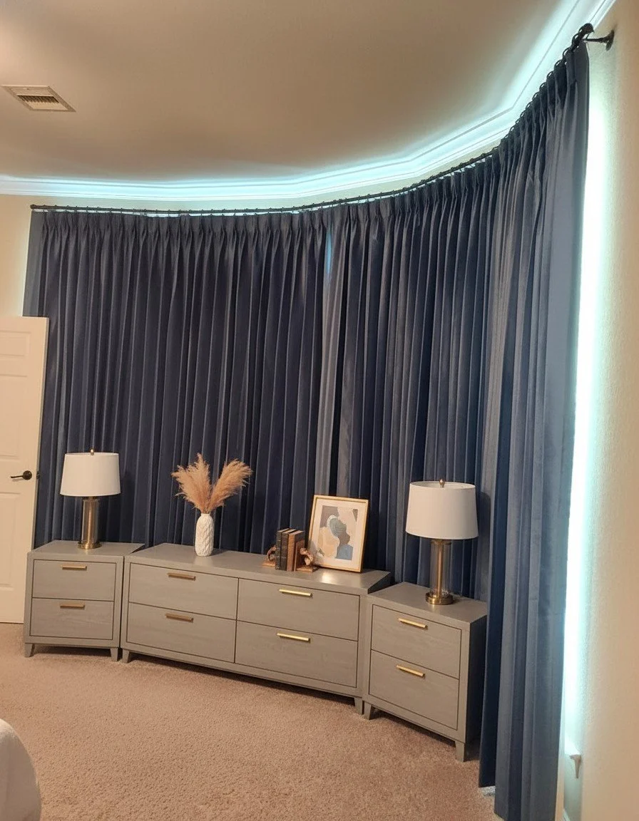 A bedroom with a grey dresser, two table lamps with white shades and gold bases, a decorative vase with pampas grass, and framed artwork, all against a large dark blue curtain covering windows, with soft lighting along the ceiling edge in Houston, TX