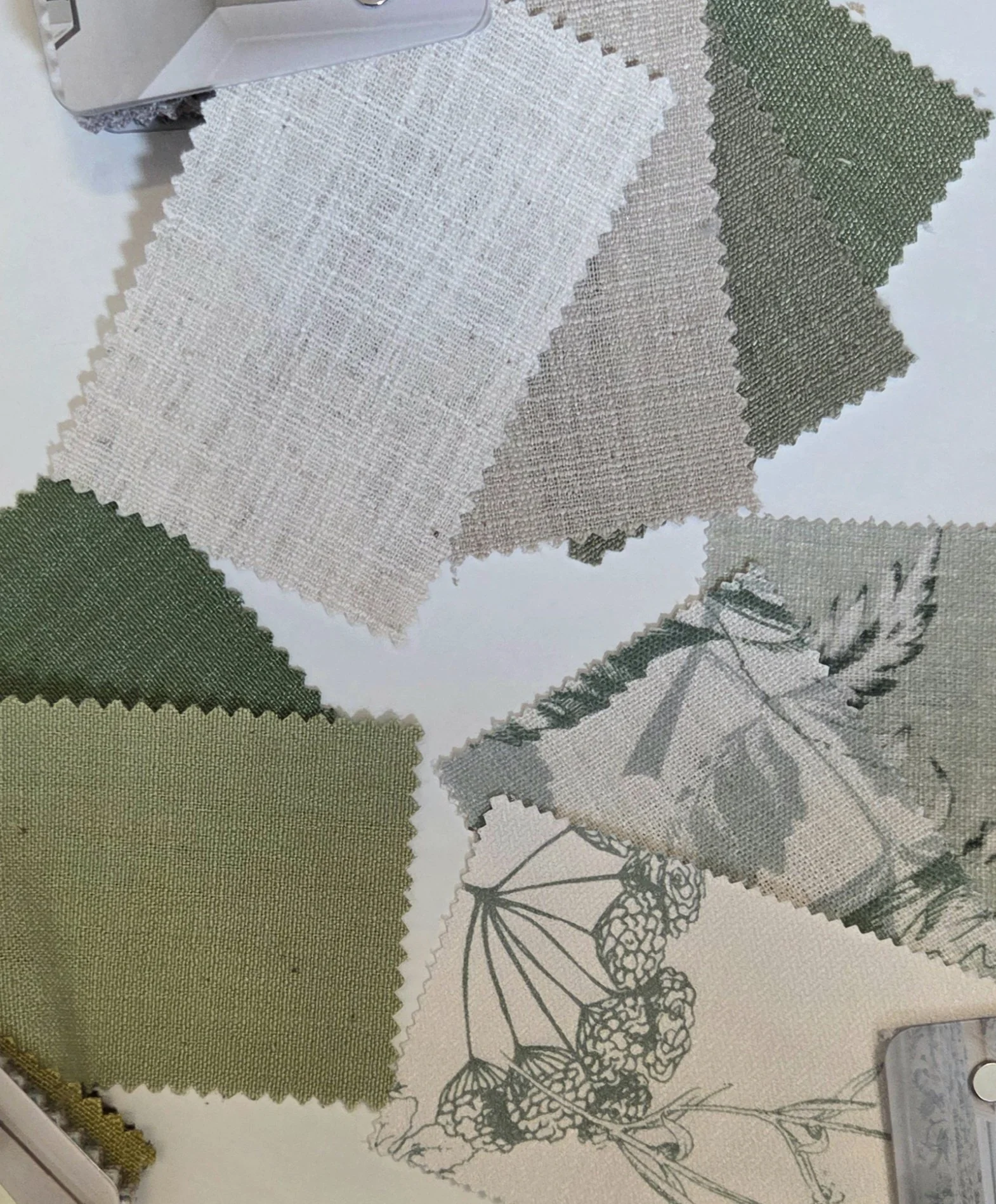 Assorted fabric swatches in neutral and green shades with textured and woven patterns, laid out on a surface next to a design sketch with a botanical illustration of flowers and leaves.