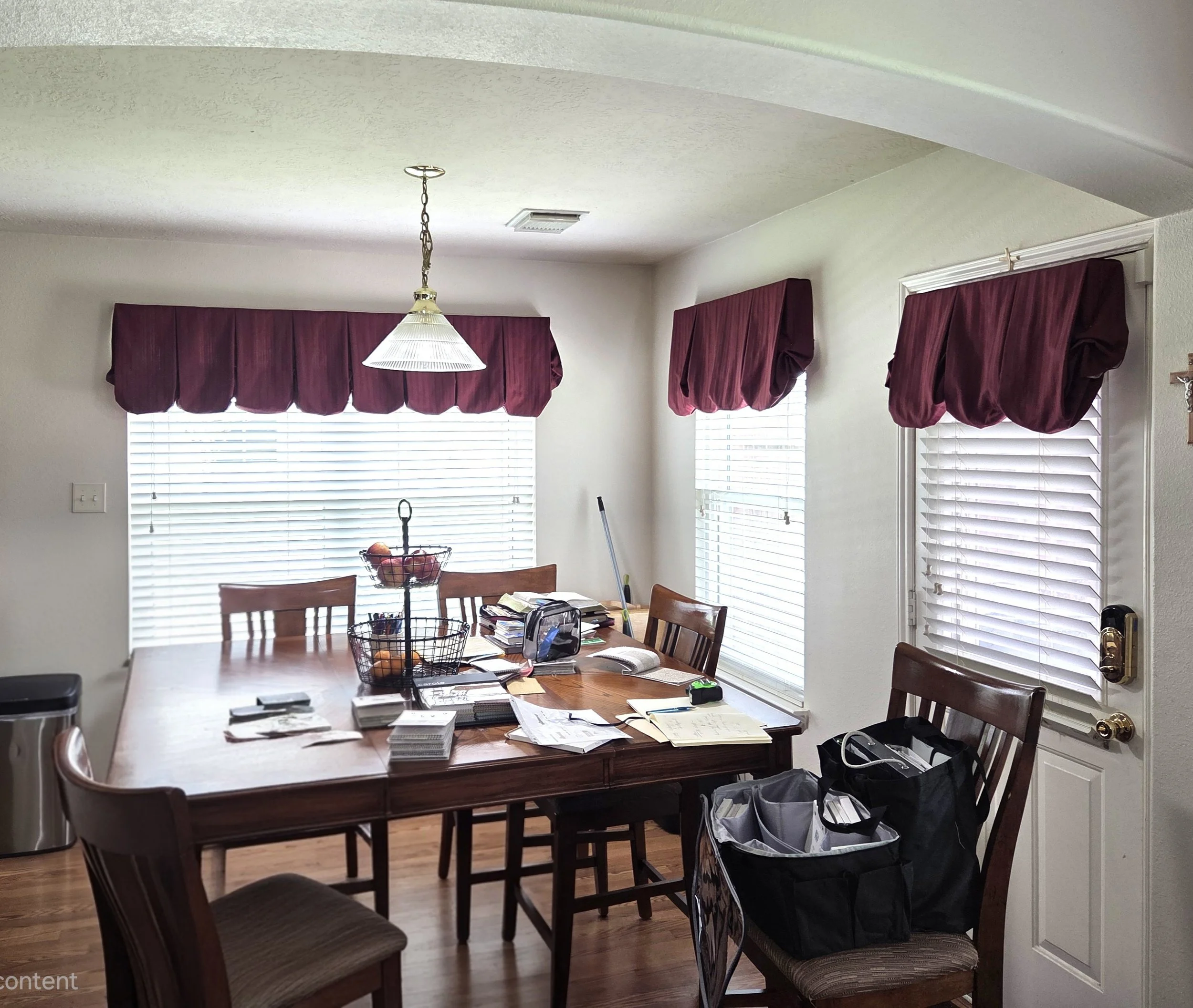 A dining room with a large wooden table, chairs, and a chandelier hanging from the ceiling. There are six windows with burgundy valances and white blinds, and the table is cluttered with papers, notebooks, and office supplies. A black bag and other items are on the floor near the door.