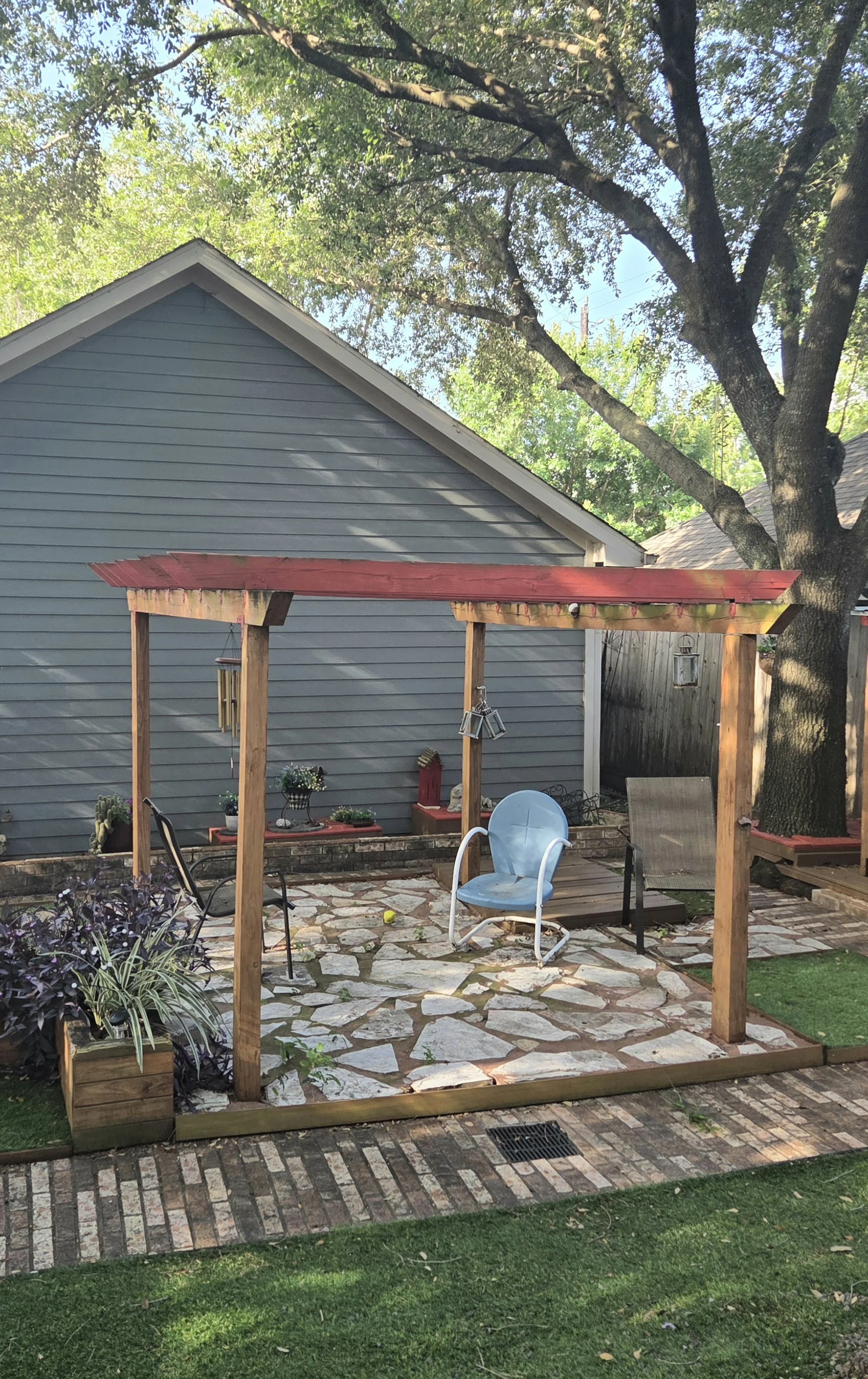 Before image of backyard patio with stone flooring, surrounded by a wooden pergola, chairs, plants, and a blue house with trees overhead Houston, TX.