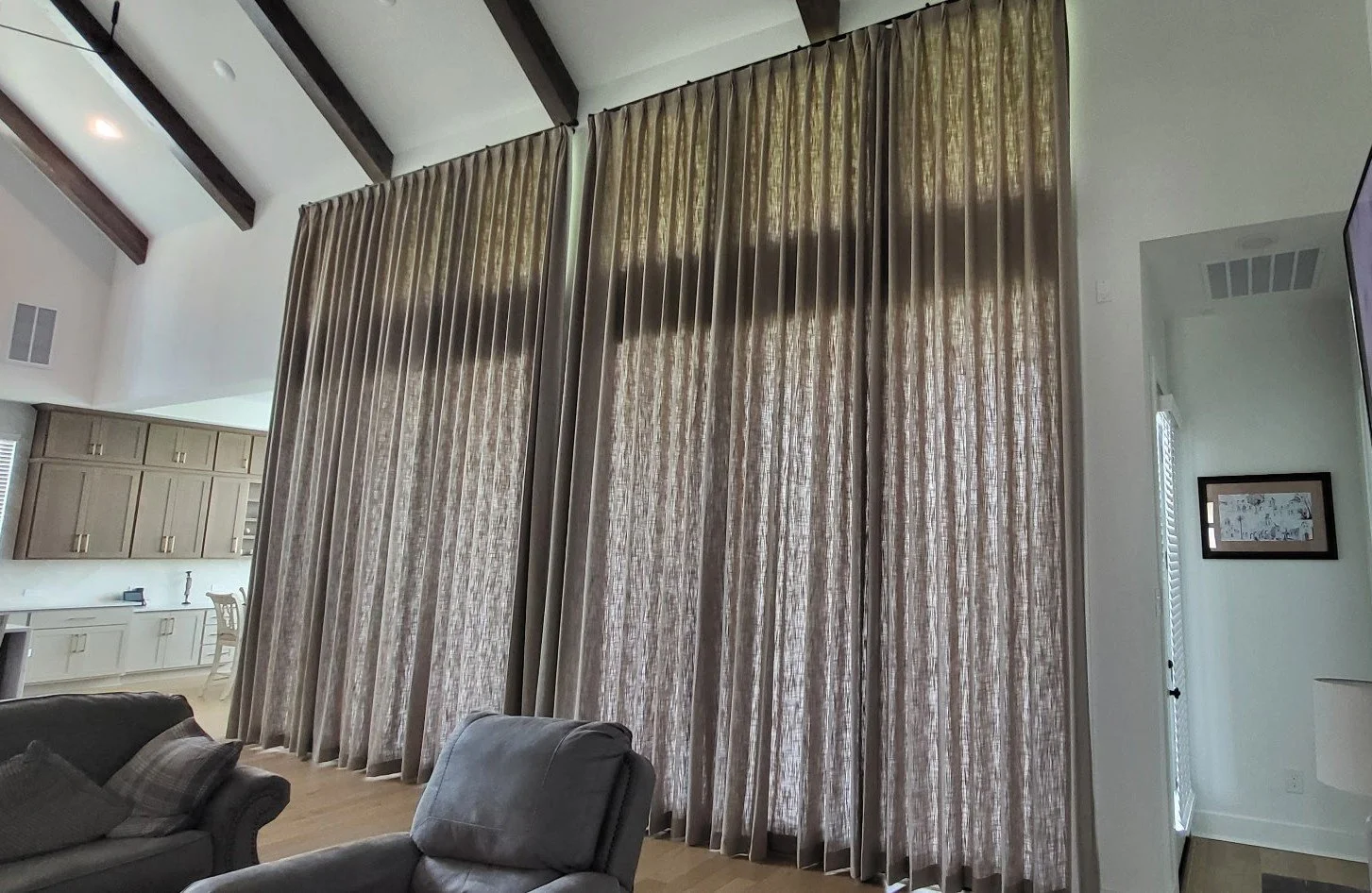 Living room with tall beige curtains covering large windows, partial view of kitchen in the background, and two gray armchairs in the foreground.