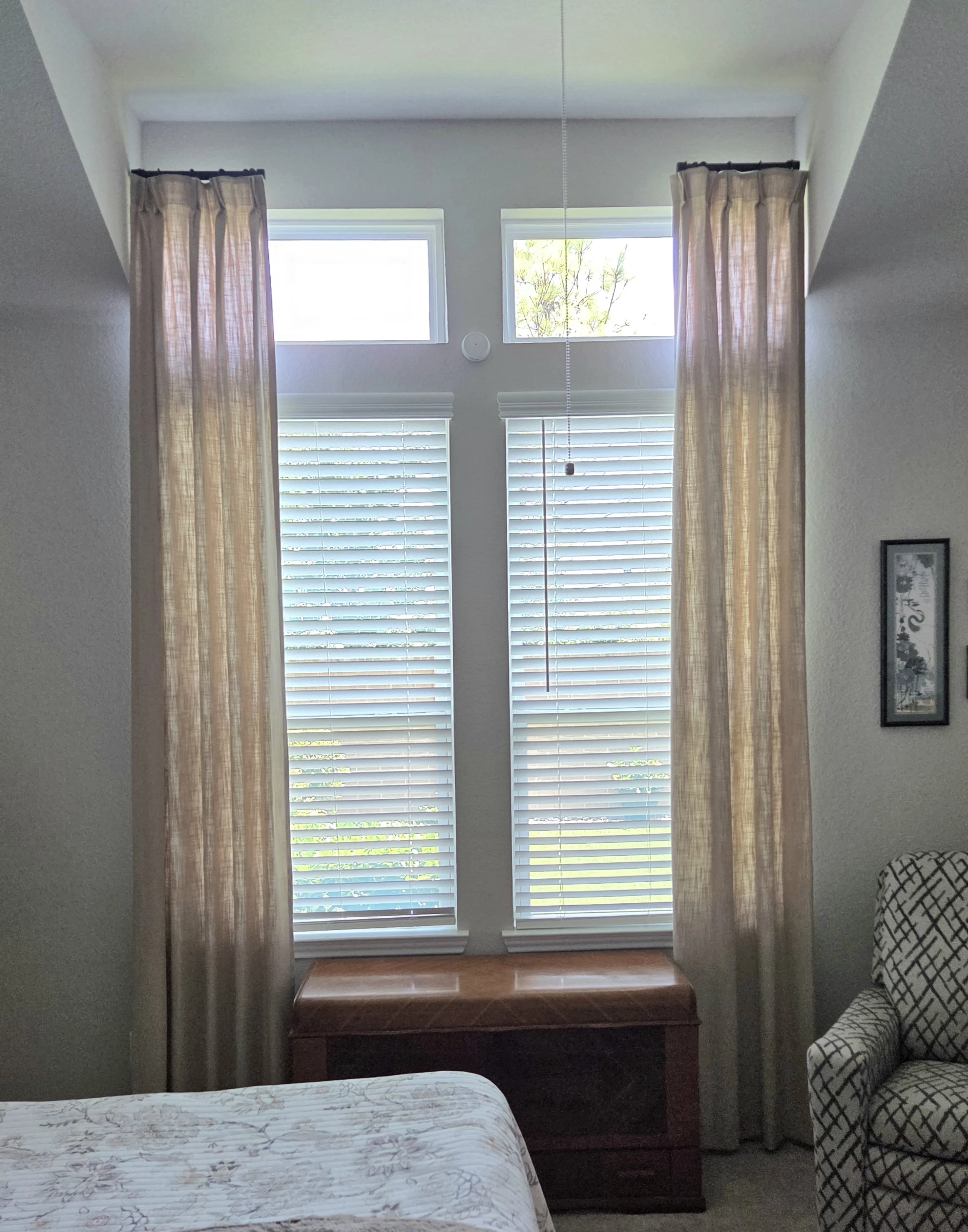 View of a bedroom with a large window blocked by custom curtains, a bench beneath the window, part of a bed in the foreground, and an armchair with a patterned cover in the right corner.