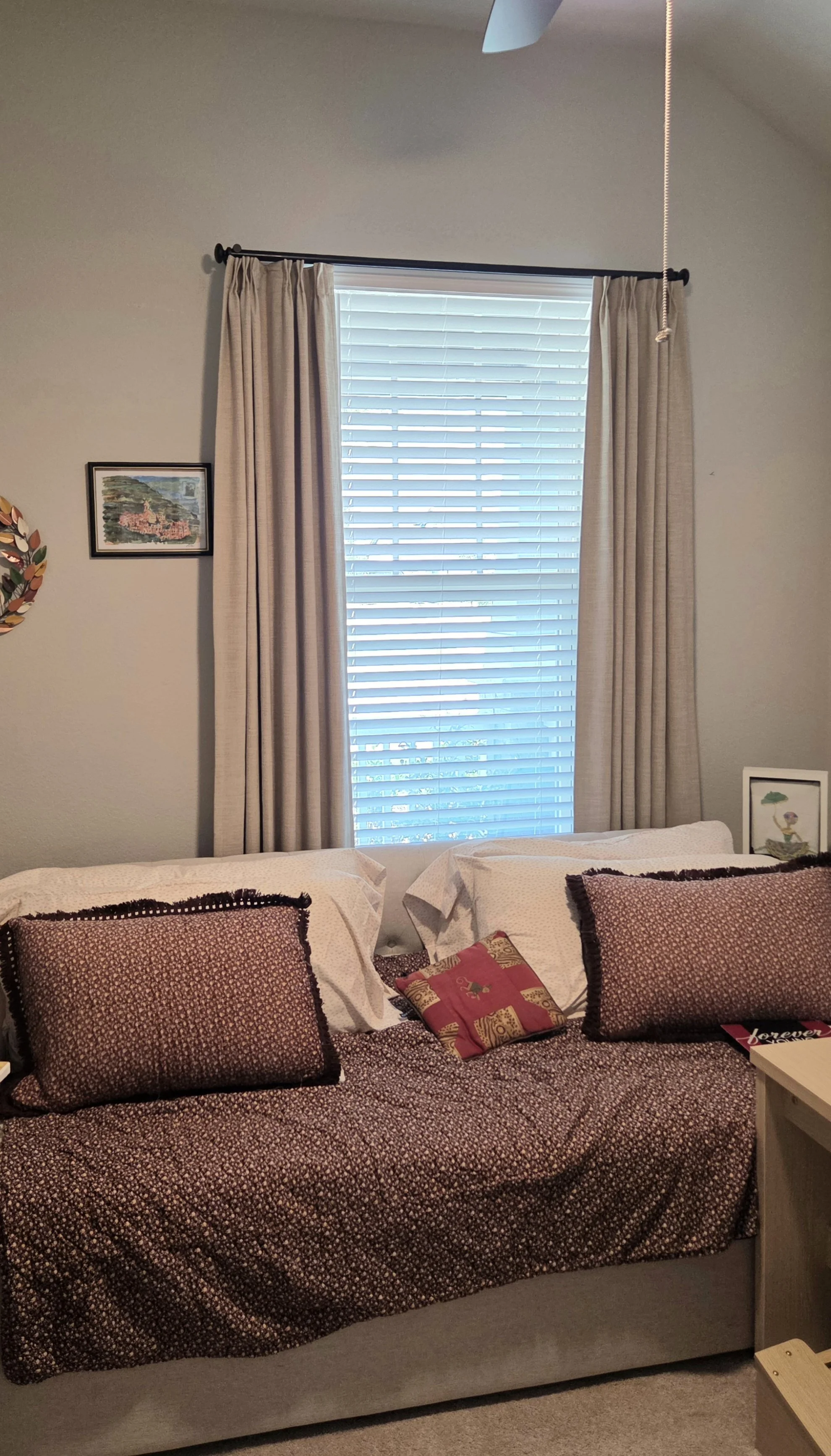A bedroom with a large window covered by white blinds and beige curtains. A bed with a patterned bedspread and multiple decorative pillows, including one with holiday-themed design. There are framed pictures on the wall and a small nightstand next to the bed.