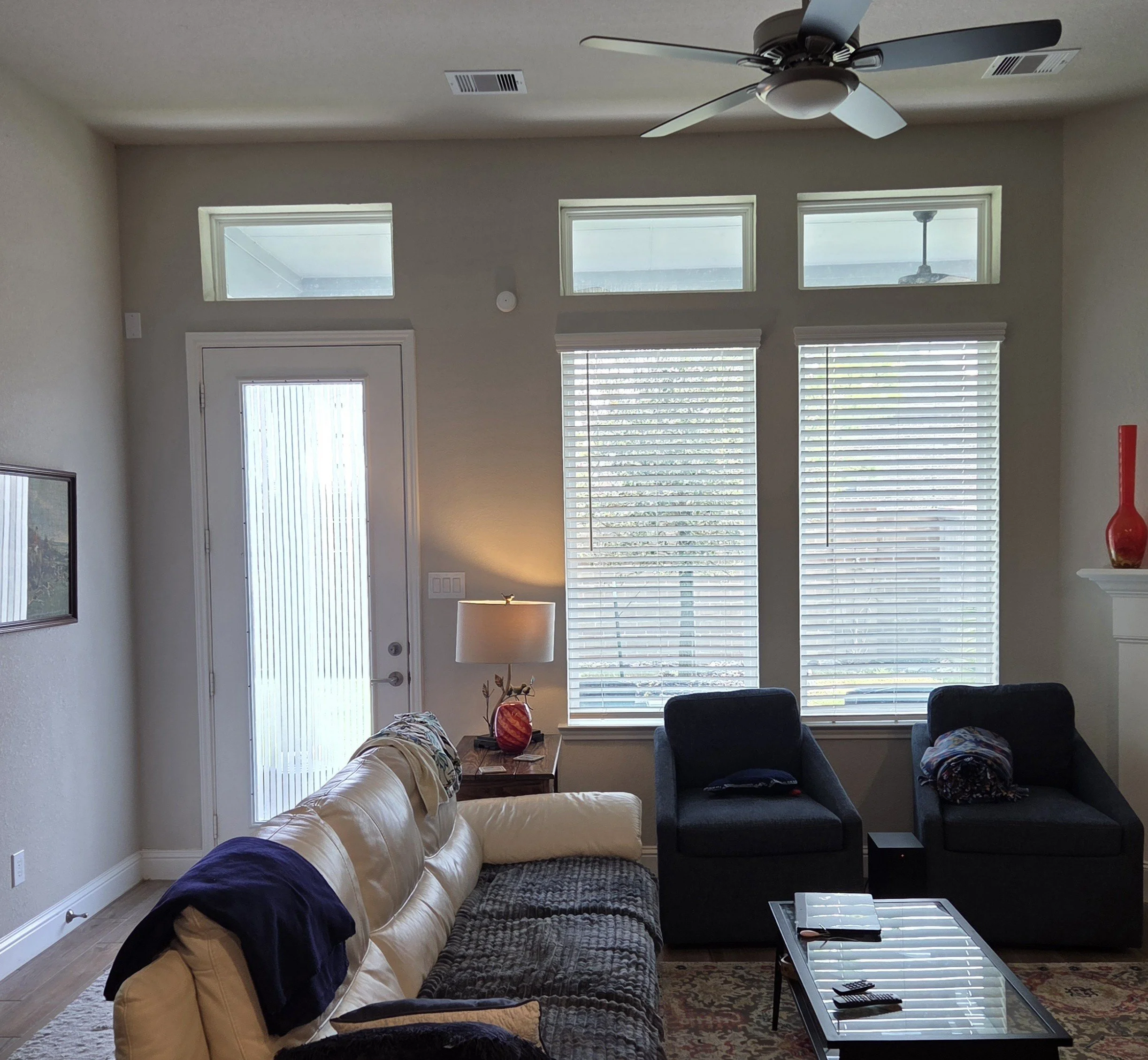 A living room with large windows and blinds, a ceiling fan, a beige sofa with pillows, a dark armchair, a coffee table, a red vase, and a lamp.