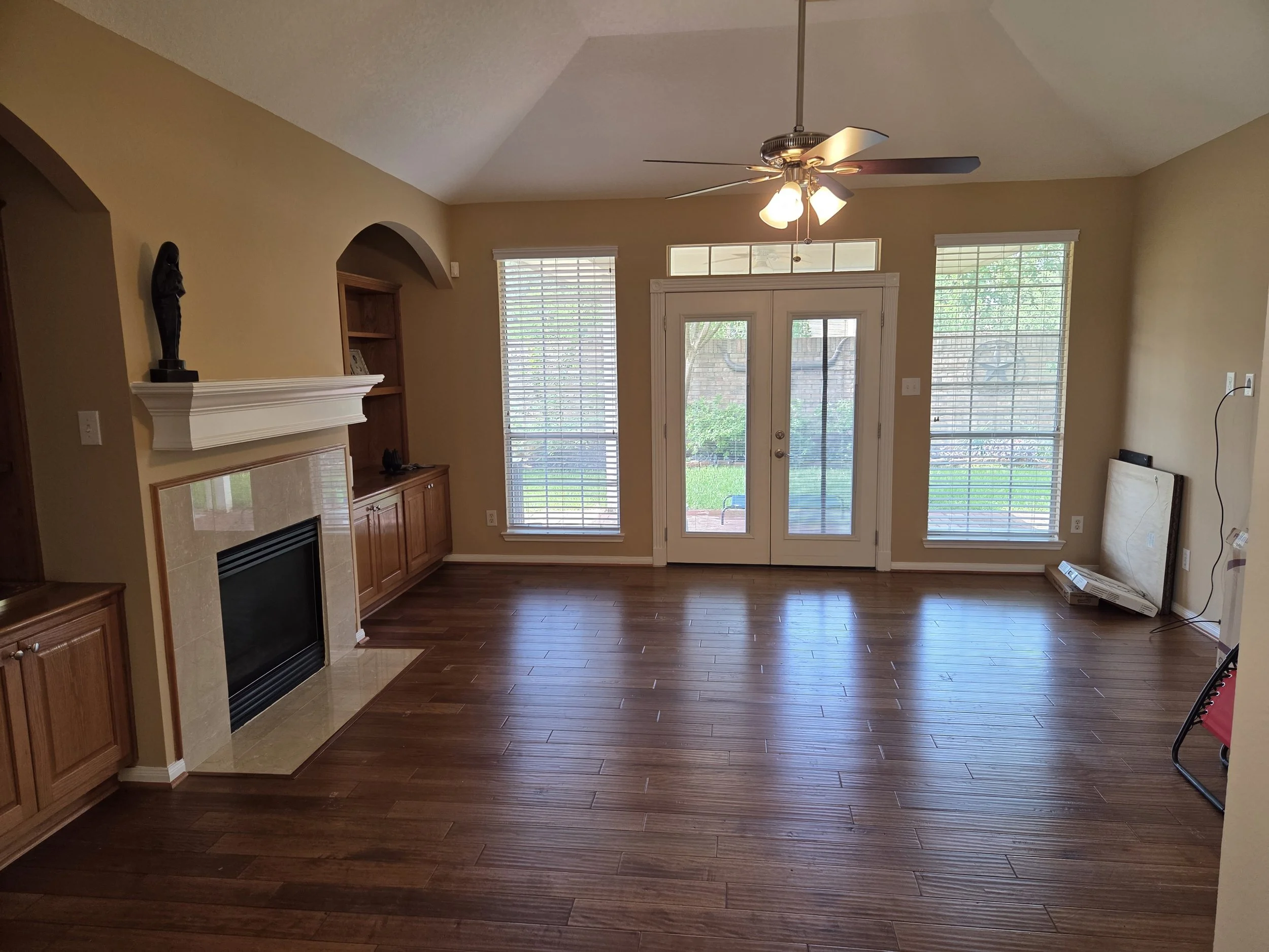 Before living room with hardwood flooring, a ceiling fan with light, large windows with blinds, a sliding glass door leading outside, a fireplace with wooden mantel, and some furniture and objects near the walls.  Houston.