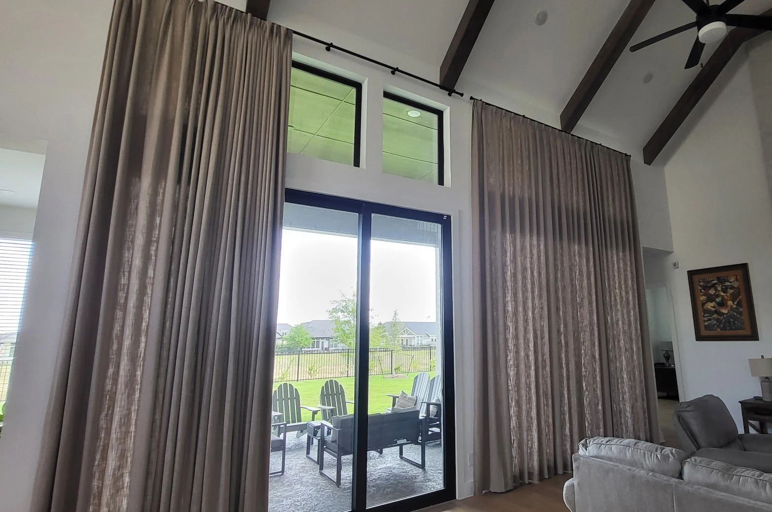 Living room with large sliding glass door and tall windows with beige curtains, opening to a patio with outdoor chairs and a view of a green backyard and neighboring houses.