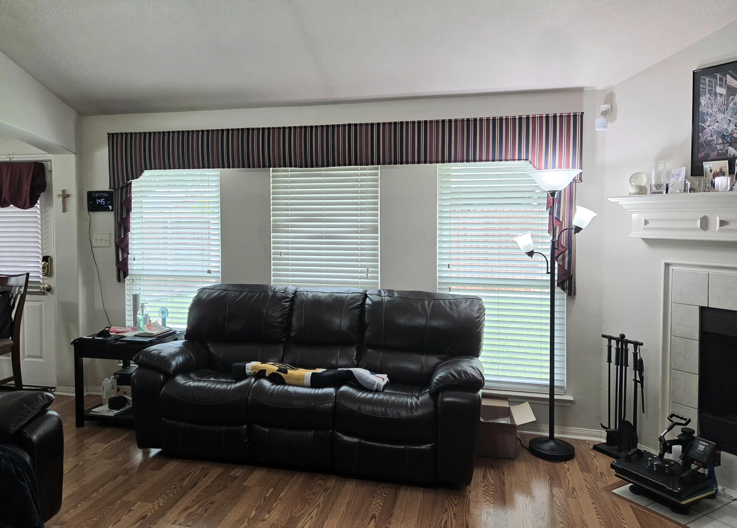 A living room with three large windows with blinds and striped valances. There is a black leather couch with a blanket on it, a small side table with hand sanitizer, and a tall floor lamp. On the right side, a white fireplace mantel with decorative items and a wall-mounted TV are visible.