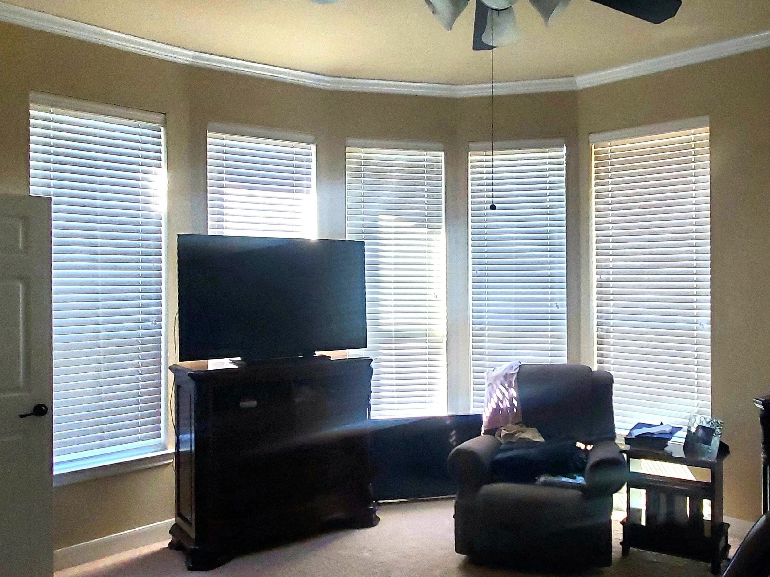 Living room with a large window wall with white blinds, a dark TV on a stand, a dark armchair with a pink cloth and some items on a side table, and light-colored walls and ceiling.