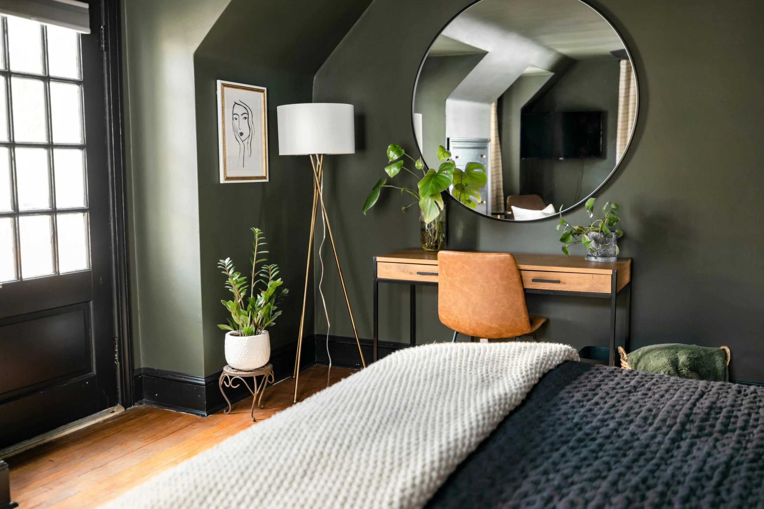 Bedroom with dark green walls, a round mirror hanging above a wooden desk with a brown leather chair, a potted plant on the desk, a white vase with green leaves, a tall white floor lamp, a framed line art picture on the wall, and a large bed with whi