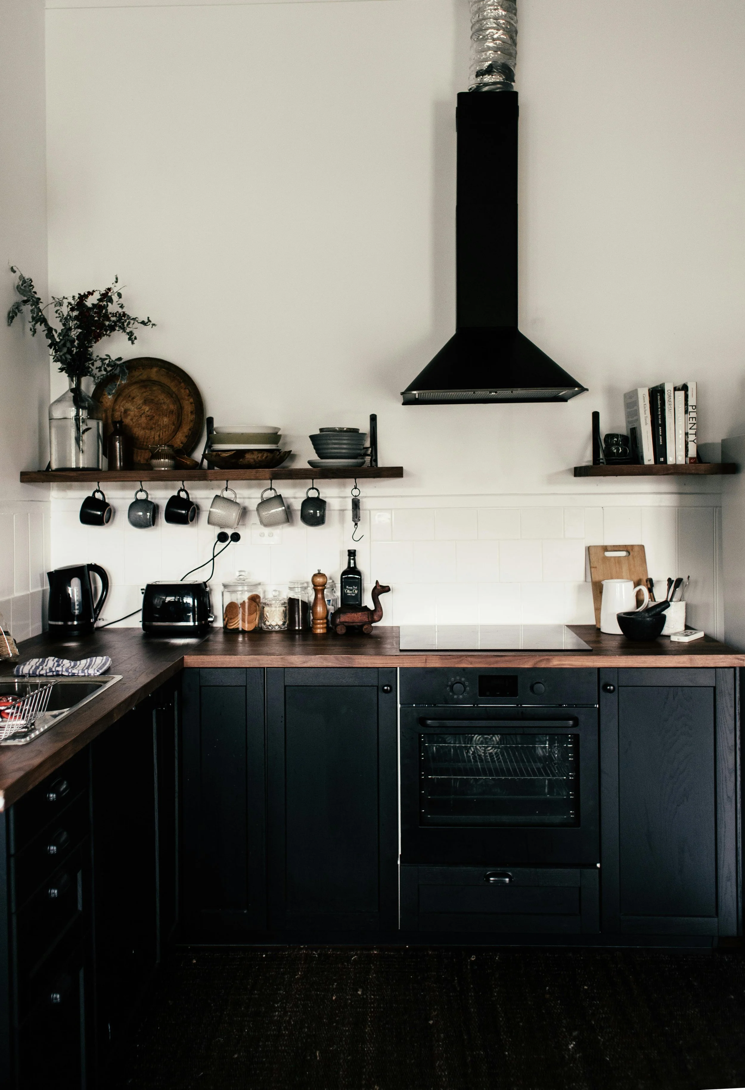 Modern kitchen with black cabinets, wooden countertop, wall-mounted shelves with dishes and books, black oven, and black chimney hood. Various kitchen appliances and utensils are on the countertop.