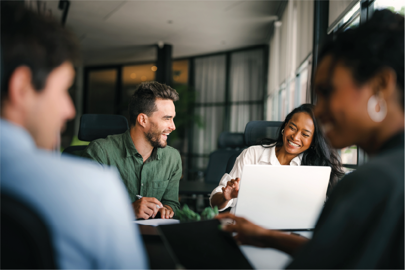 Four people sitting around a table in a modern office, smiling and having a conversation with a laptop open in front of them.