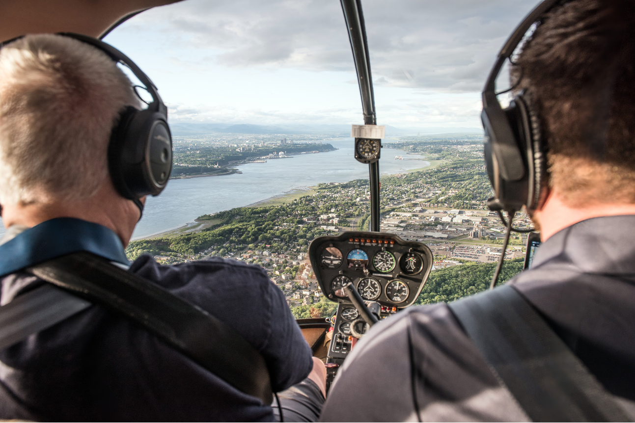 Deux hommes pilotant un hélicoptère survolant une ville avec une rivière et des montagnes en arrière-plan.