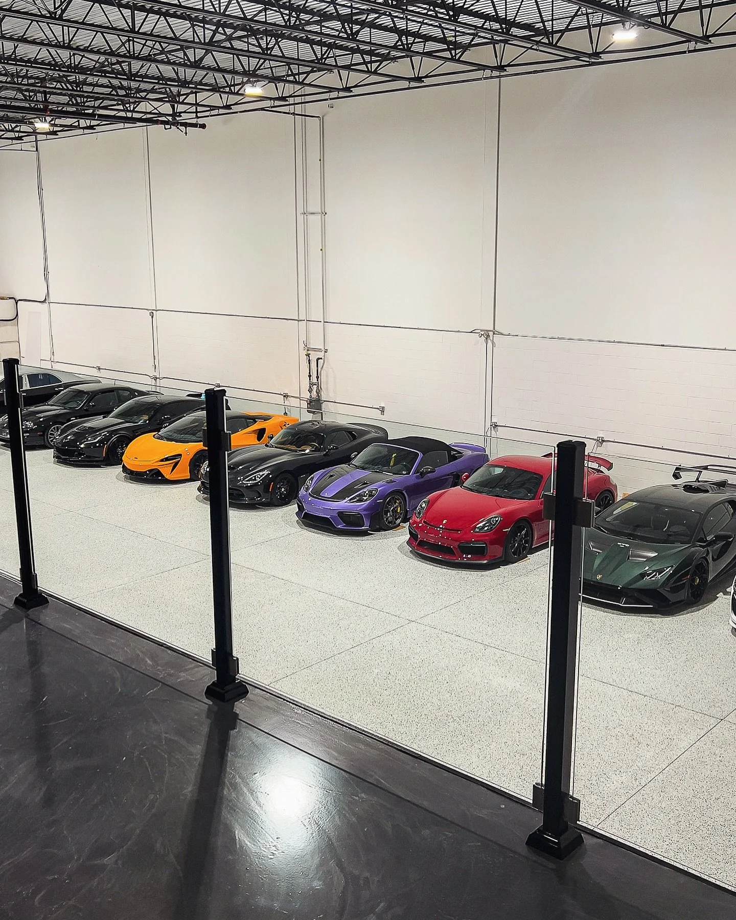 Une rangée de voitures de sport luxueuses dans un entrepôt, comprenant une Lamborghini jaune, une Ferrari rouge, des Porsche violettes et vertes, ainsi que d'autres voitures de haute gamme.