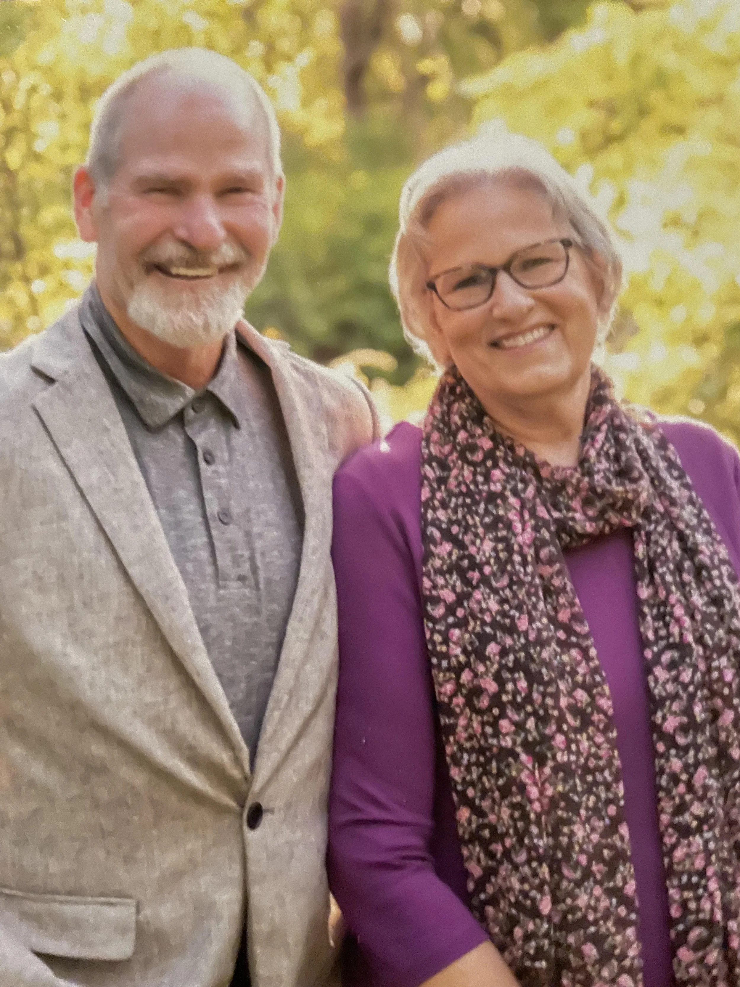 A smiling older man with a beard and a smiling older woman with glasses, standing outdoors with yellow autumn leaves in the background.