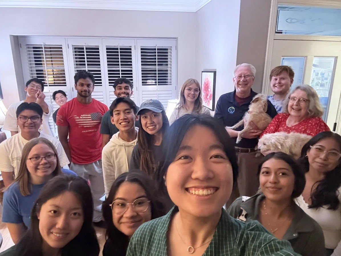 Group of people indoors taking a selfie, smiling, with some holding dogs, in a well-lit living room with large windows and shutters.
