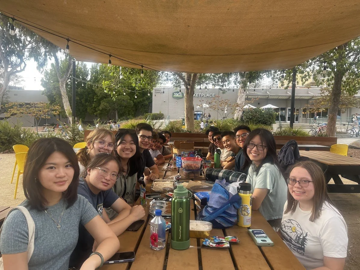 Group of people sitting at a picnic table outdoors under a canopy, enjoying a meal and fellowship.