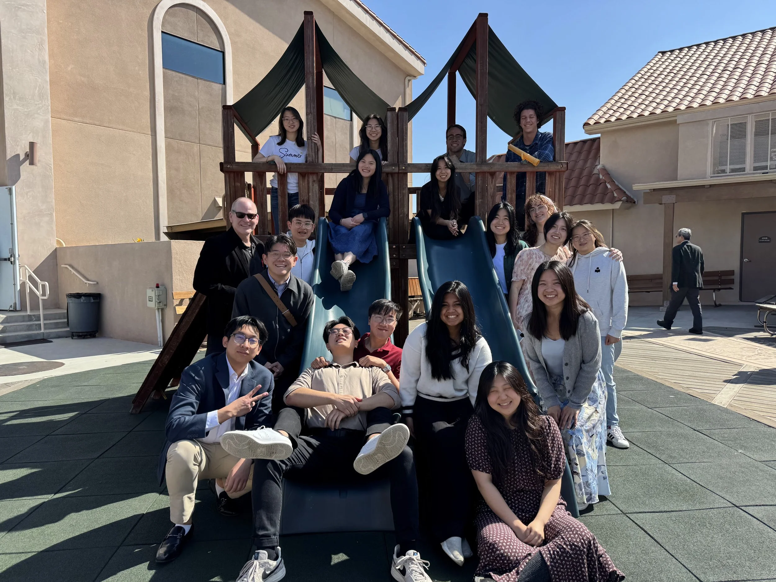 A group of smiling people gathered on a playground slide and surrounding area, posing for a photo outside on a sunny day with buildings in the background.