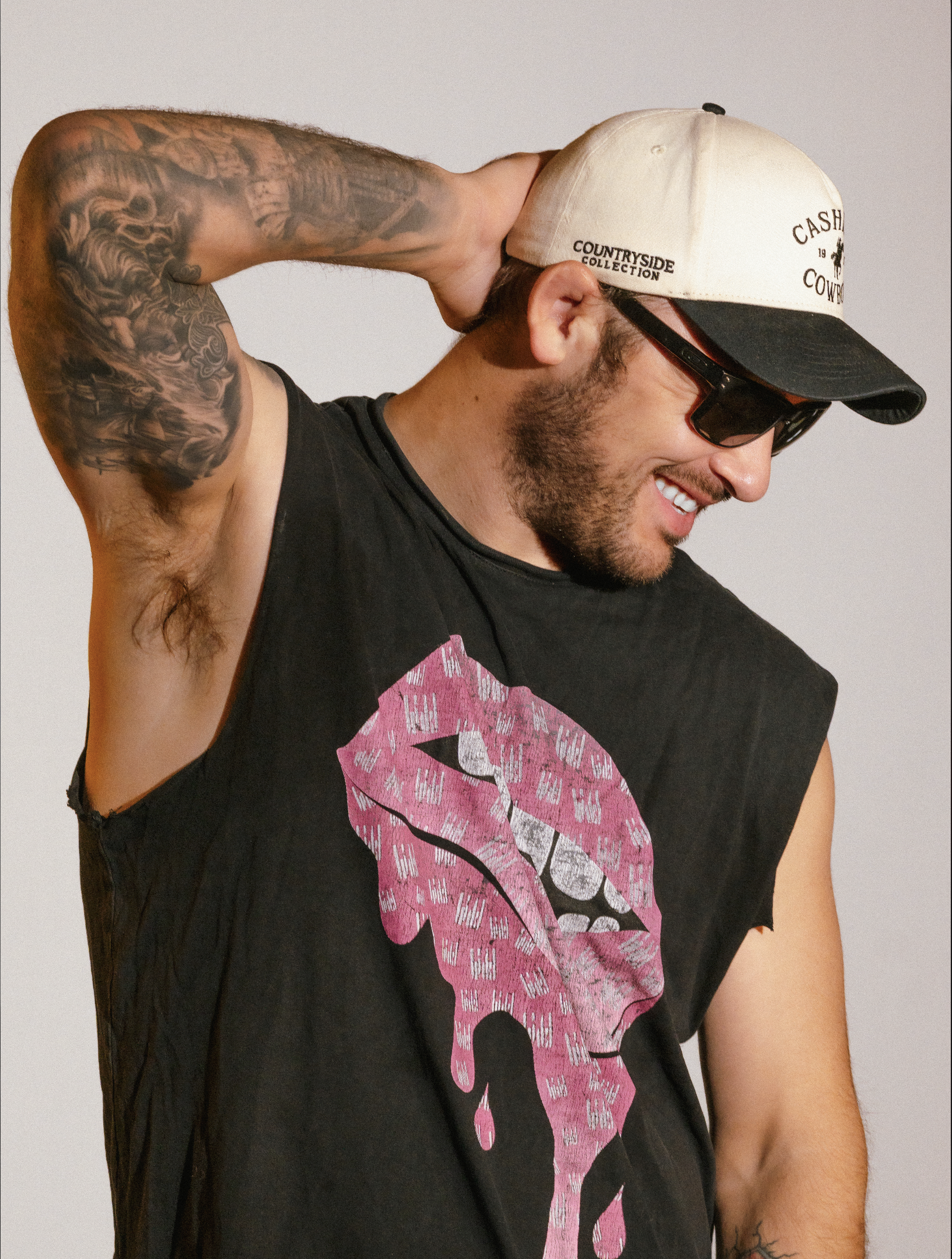 Casual lifestyle portrait of a tattooed man wearing a hat and sunglasses, captured with crisp lighting on a white studio backdrop.