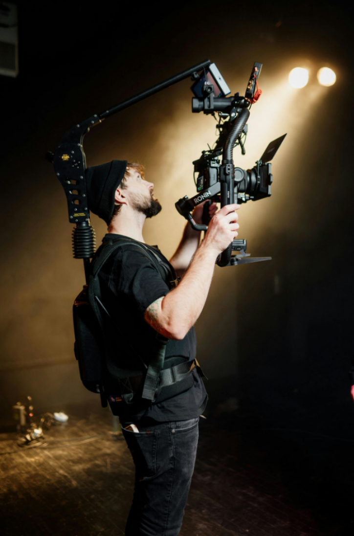 A professional cinematographer in a dark studio using a heavy-duty camera rig and gimbal under cinematic lighting to capture high-production commercial visuals.