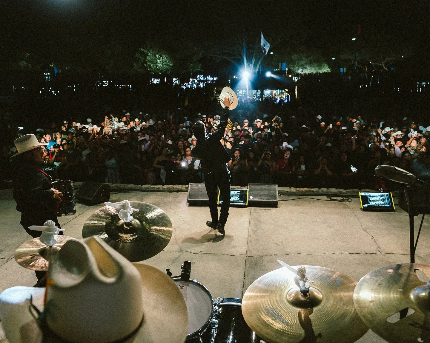 Professional event coverage from the stage of a live outdoor music concert, showing the performer raising a hat to a massive crowd.