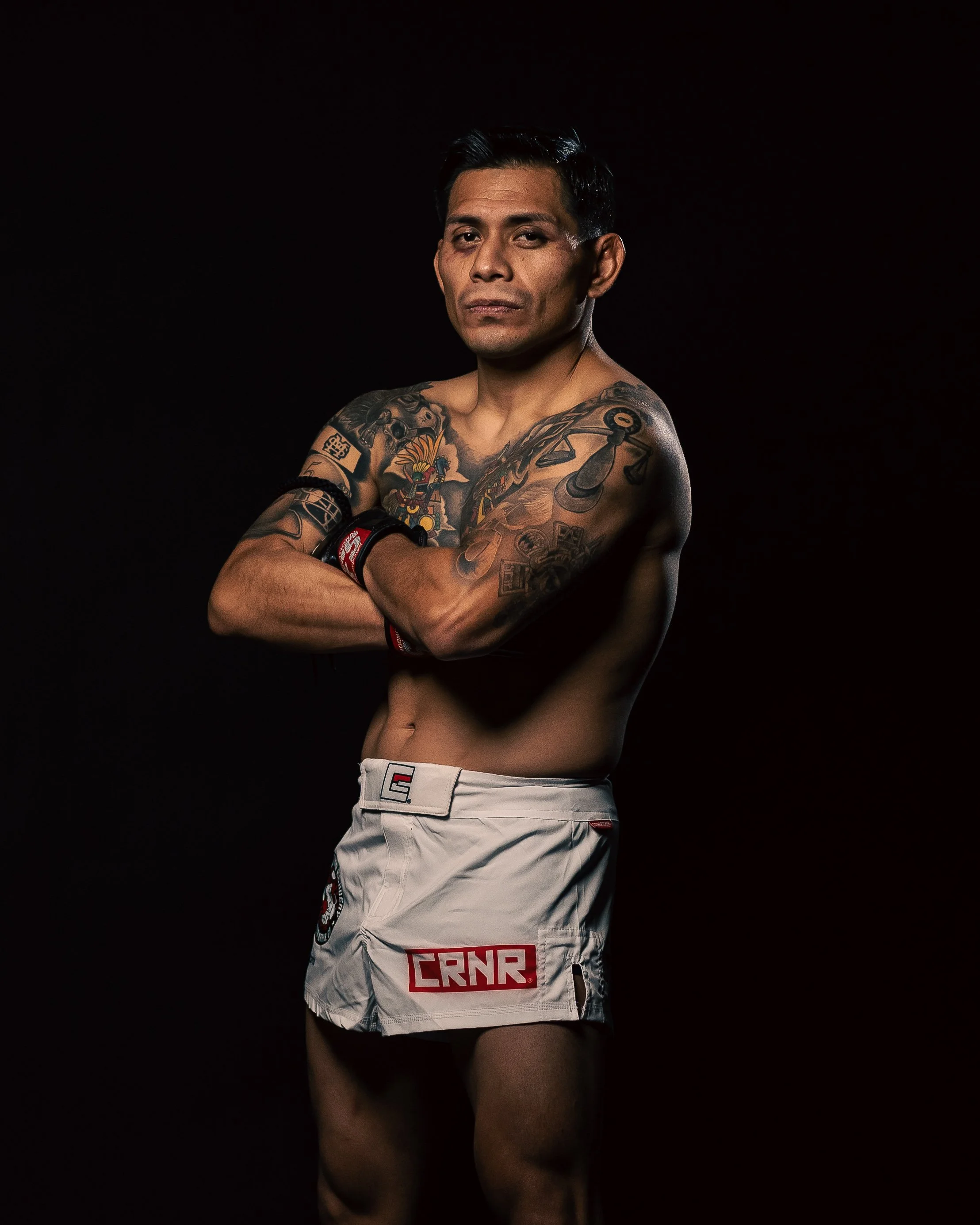 Dramatic fitness portrait of a tattooed man with arms crossed, utilizing professional dark set lighting for high-contrast photography.