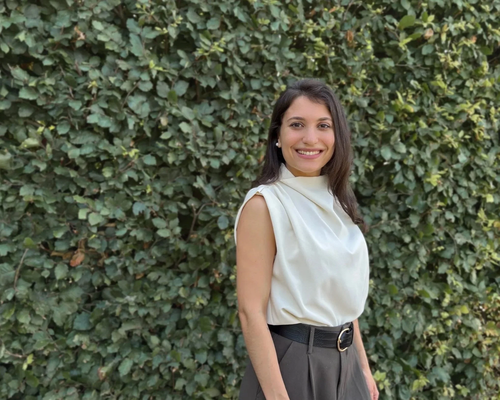 A woman smiling outdoors in front of a large green leafy hedge, wearing a sleeveless white top and dark trousers with a black belt.